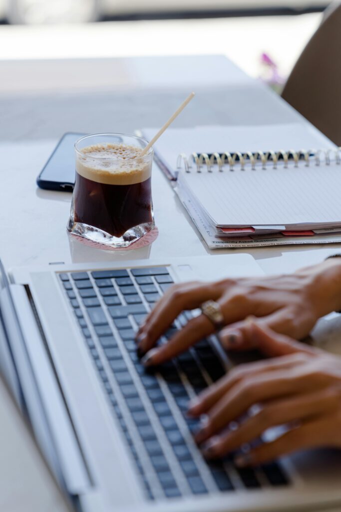 Hands typing on a laptop with iced coffee and notebook nearby, creating a productive work environment.